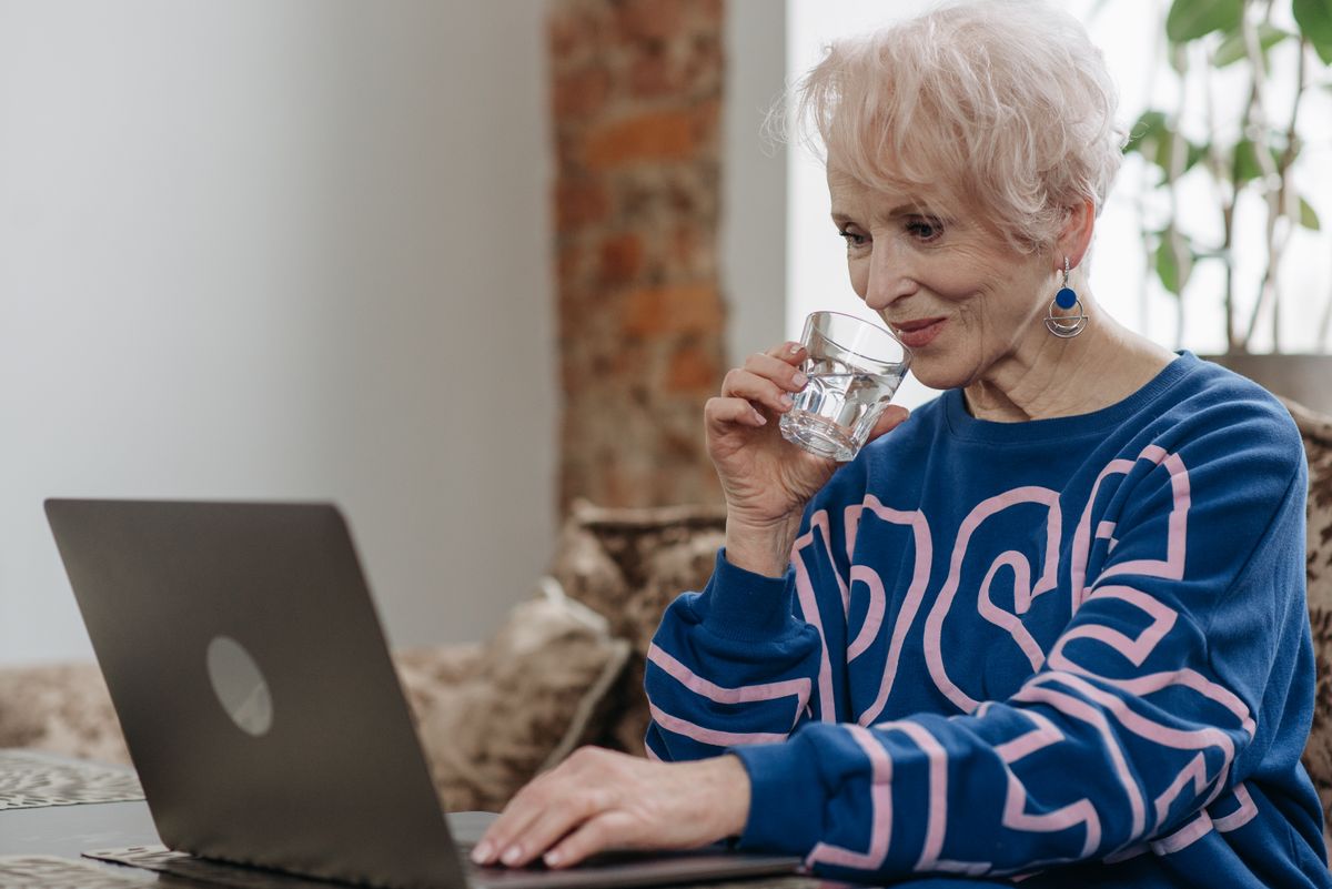 Older woman sipping a glass of water at her laptop, easing menopause dry mouth and bad breath